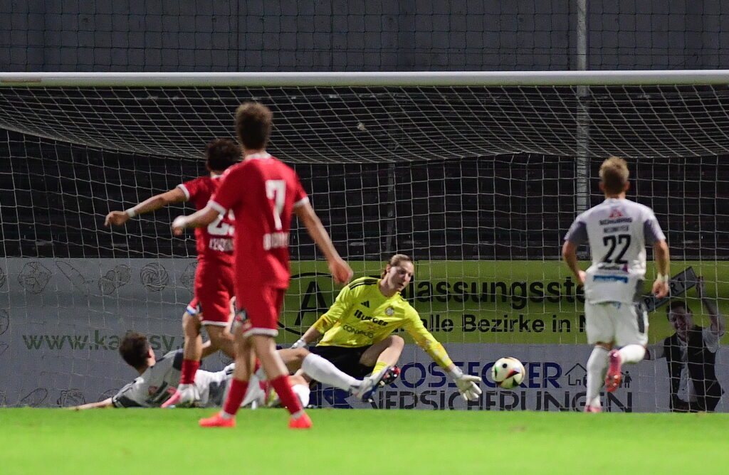 Fußball Torwart im gelben Dress pariert Ball vor Spielern in roten Trikots im Stadion.